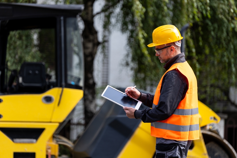 Construction worker wearing a hard hat and safety vest inspecting heavy equipment while documenting information on a tablet.